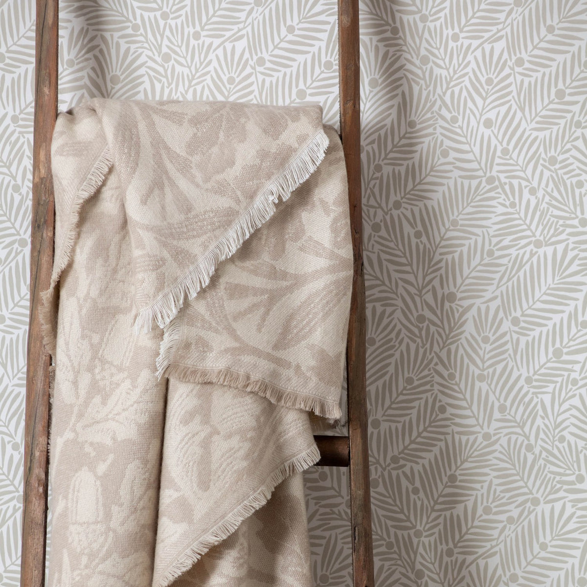 Acorn Tonal Jacquard Linen Throw in beige, draped over a wooden ladder with fringed edges, showing a woven leaf and acorn pattern against leaf-print wallpaper.