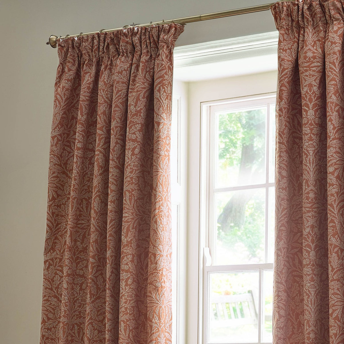 Acorn Tonal Jacquard pencil pleat curtains in brick, woven acorn branch pattern in pale cream, framing a window in a light-toned room.