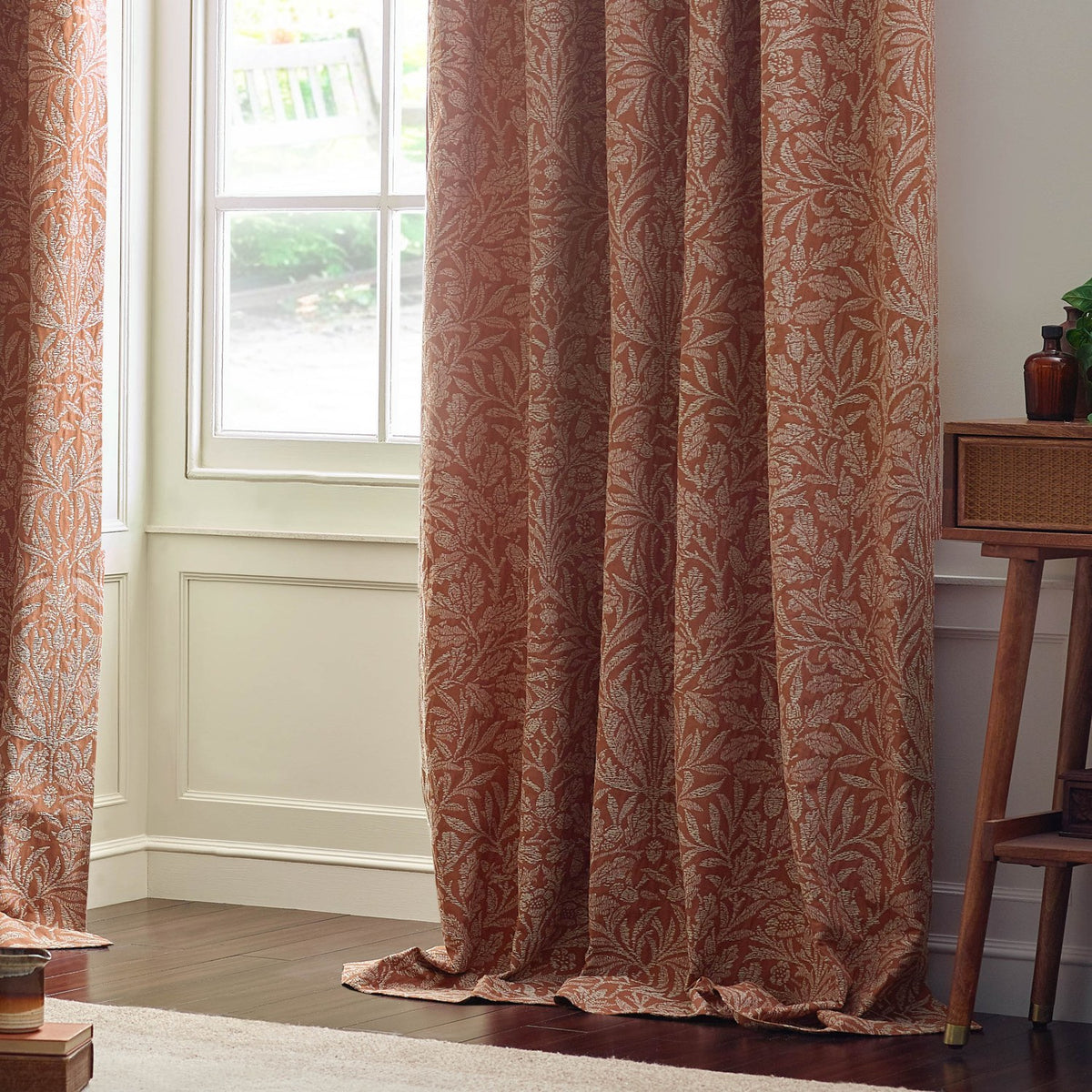 Acorn Tonal Jacquard pencil pleat curtains in brick, showing a tonal acorn branch pattern in pale cream on rust fabric, styled floor-length beside a bay window.
