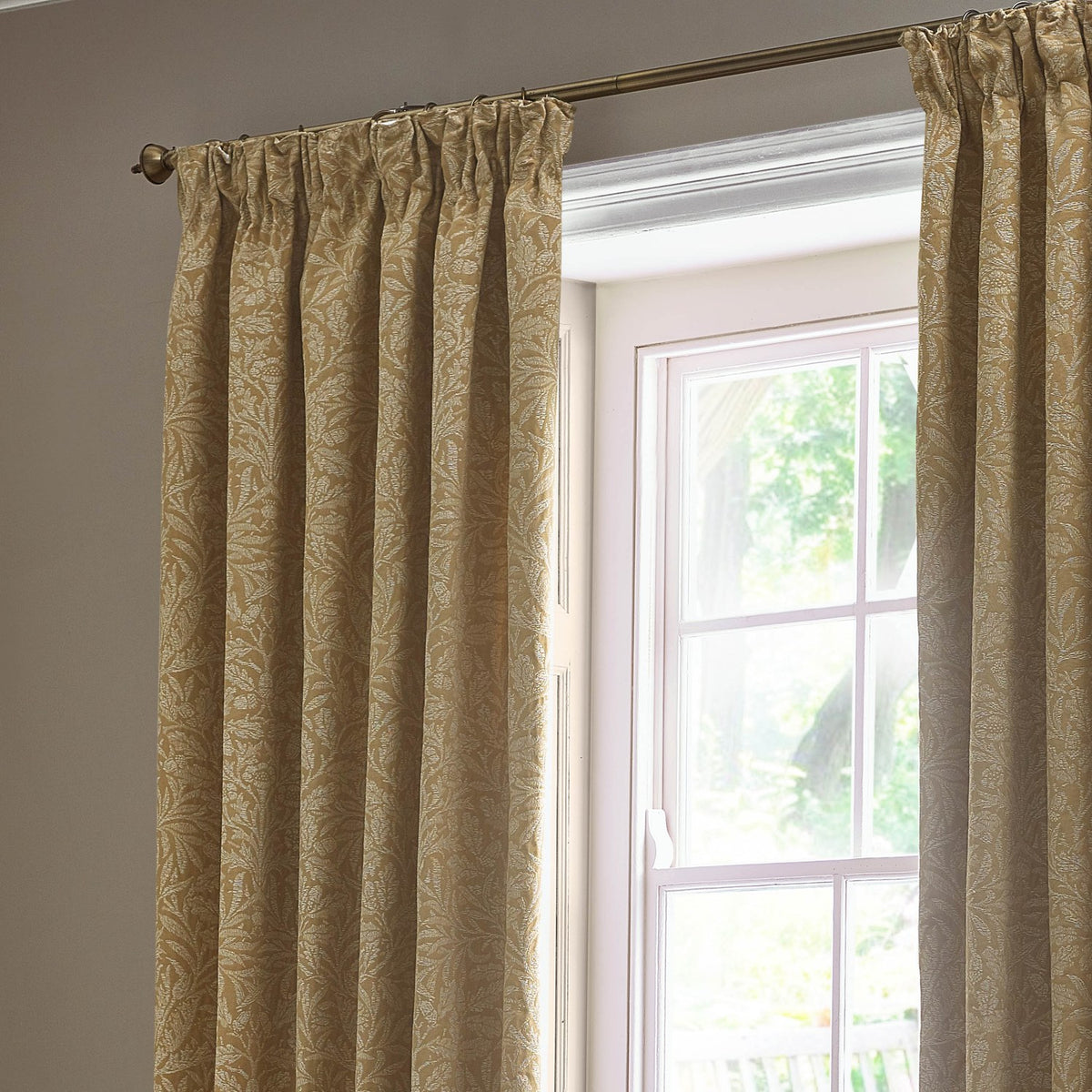 Pencil pleat curtains in saffron-gold jacquard with a tonal Acorn leaf design, shown pleated on a bronze pole beside a bright window.