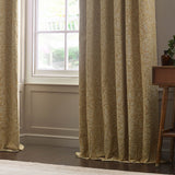 Saffron-gold pencil pleat curtains with tonal Acorn jacquard pattern, shown draped by a bay window in a living room with a wooden side table.