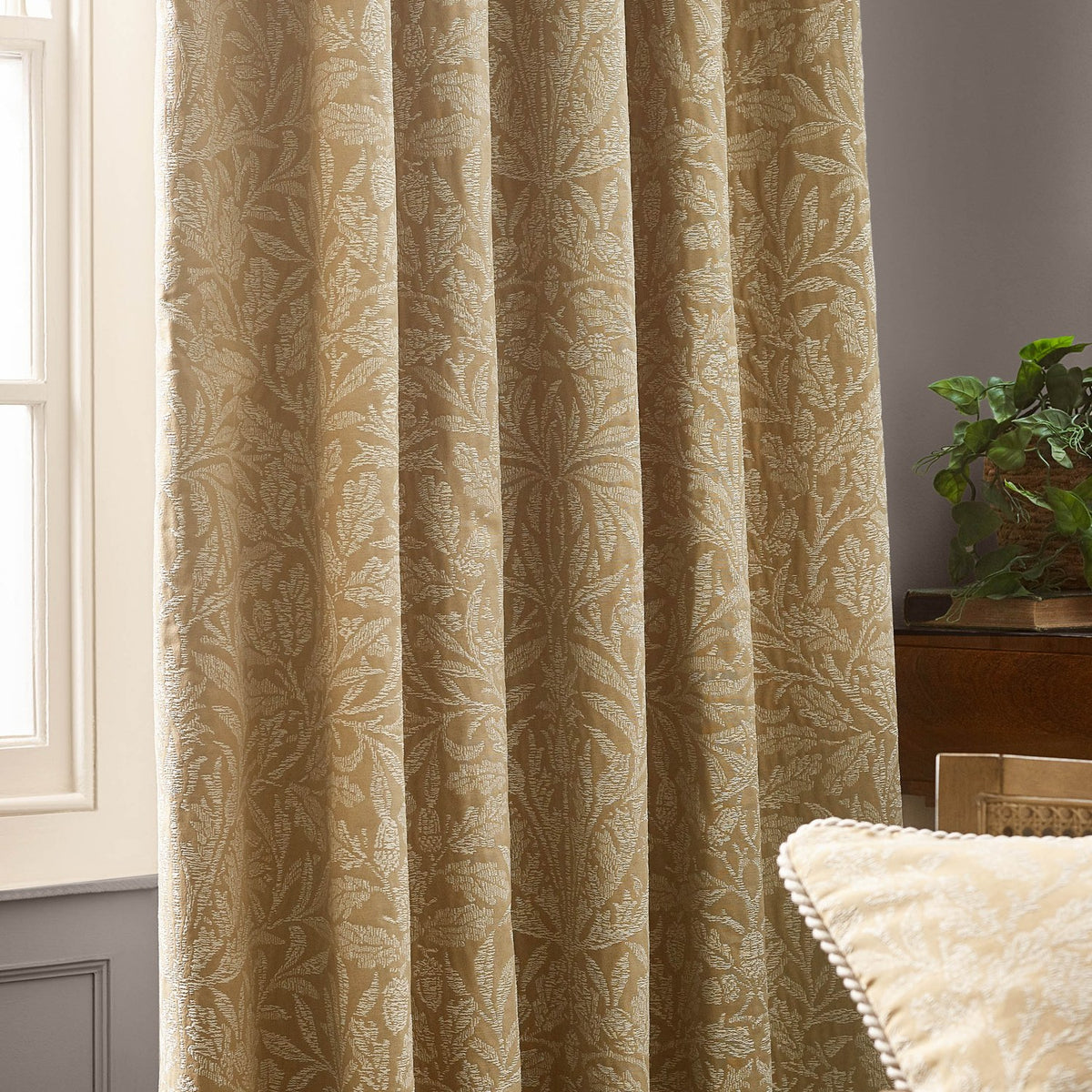 Pencil pleat curtains in saffron-gold tonal jacquard with the Acorn leaf and branch pattern, shown hanging in folds beside a window in a living room setting.