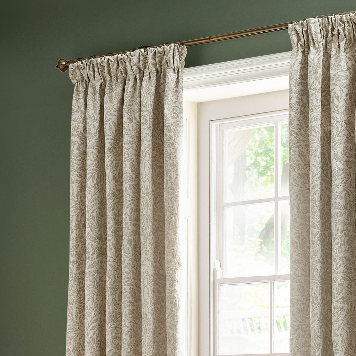 Acorn Tonal Jacquard pencil pleat curtains in cream Willow, shown with pencil pleat heading on a brass pole beside a window in a green-walled room.