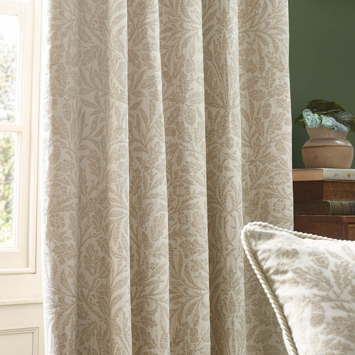 Acorn Tonal Jacquard pencil pleat curtains in Willow, cream fabric with woven acorn and oak leaf pattern, shown hanging in a living room beside a matching cushion.
