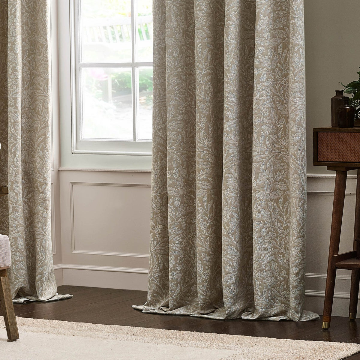 Acorn Tonal Jacquard Linen pencil pleat curtains in beige with woven acorn and oak leaf pattern, shown pooling on the floor beside a window in a living room.