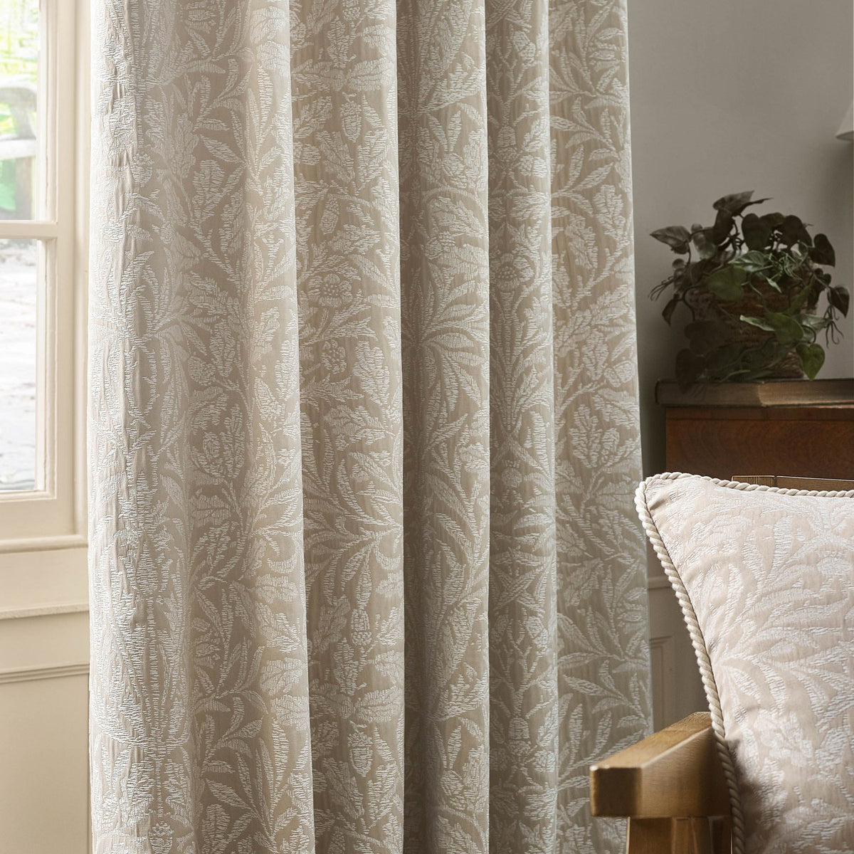 Acorn Tonal Jacquard Linen pencil pleat curtains in beige, woven with an acorn and oak leaf pattern, shown pleated at a window beside a cushion and side table.