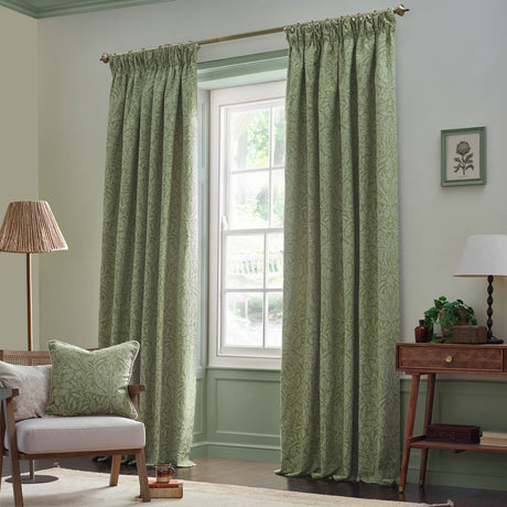 Sage green pencil pleat curtains with tonal Acorn jacquard leaf pattern, shown hanging on a brass pole in a living room window setting.