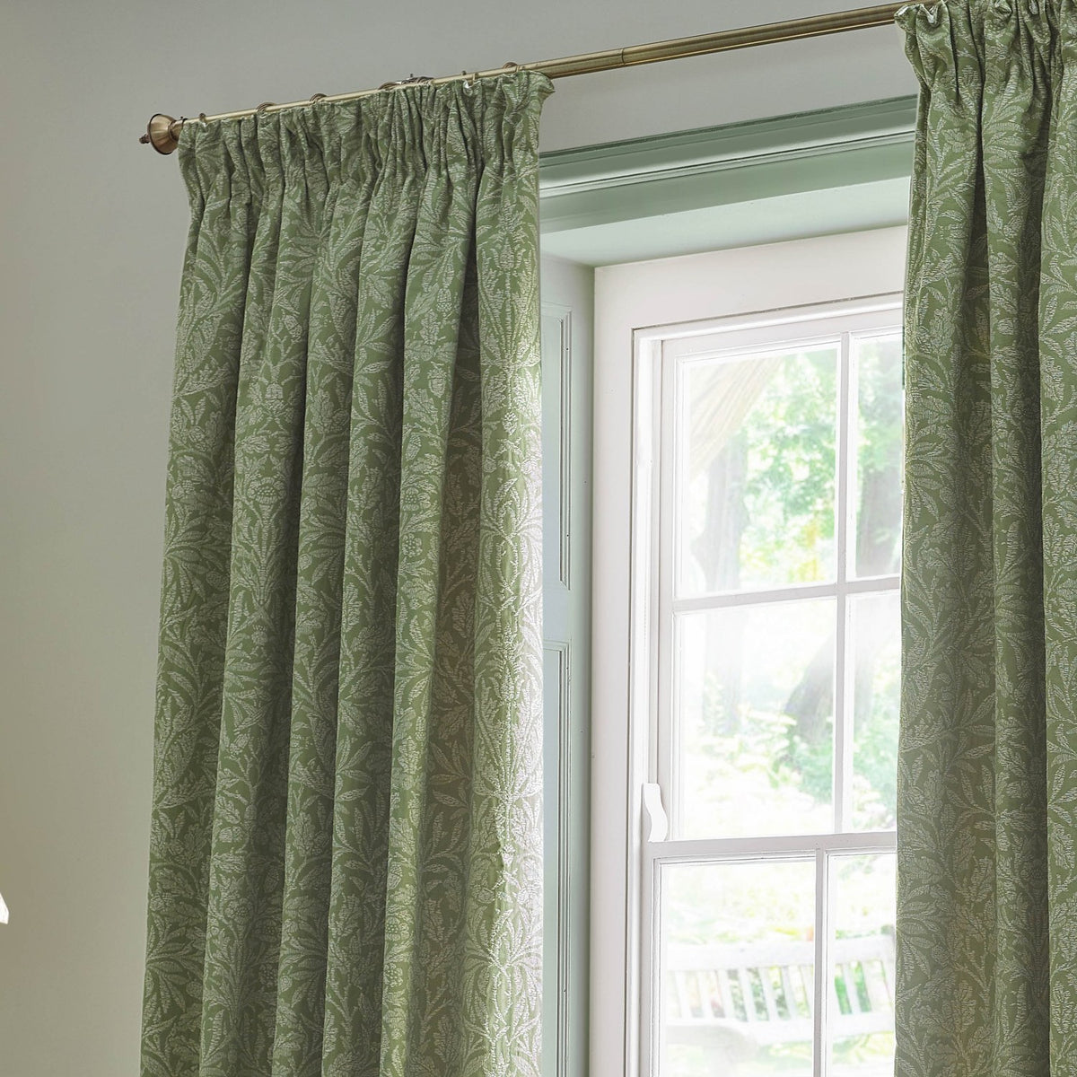 Sage green pencil pleat curtains with a tonal Acorn foliage jacquard pattern, hung from a brass pole beside a white-framed window.