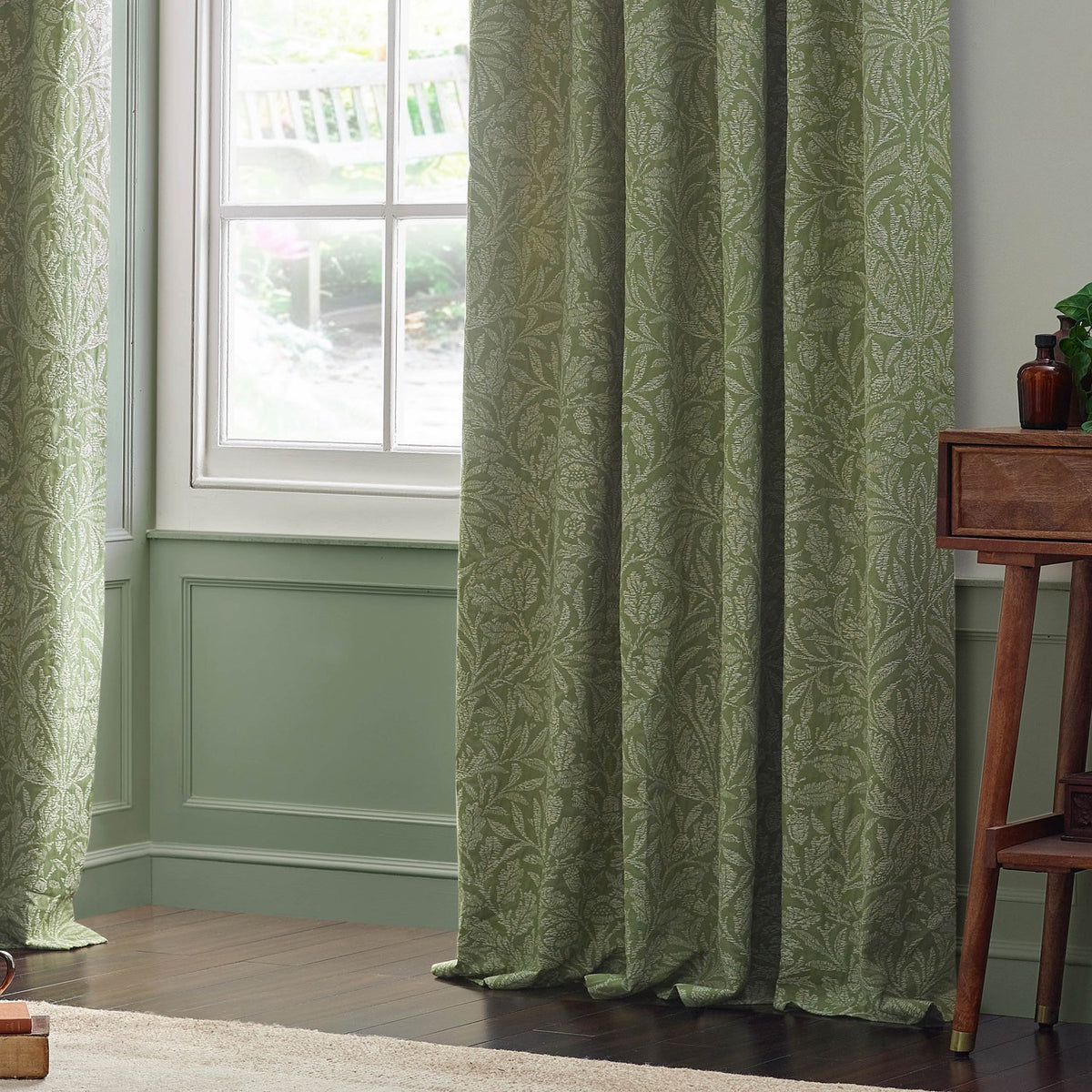 Sage green pencil pleat curtains in the Acorn tonal jacquard design, shown hanging beside a window in a living room with panelled walls and a dark wood side table.
