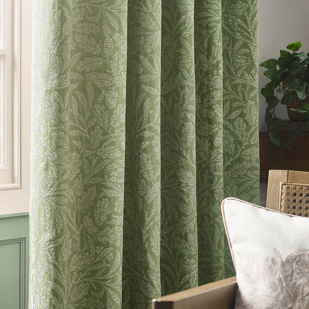Acorn Tonal Jacquard pencil pleat curtains in sage, showing woven oak leaf and acorn pattern in soft two-tone fabric, hung in a living room beside a cushioned chair and potted plant.