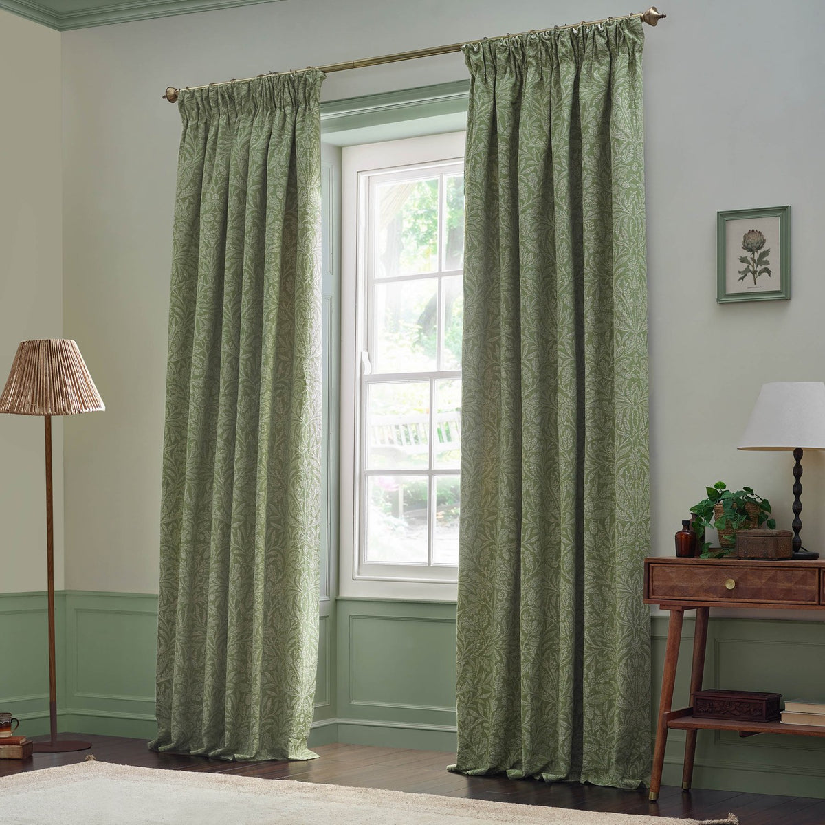 Sage green pencil pleat curtains with a tonal Acorn jacquard leaf pattern, shown framing a window in a living room with pale green walls.