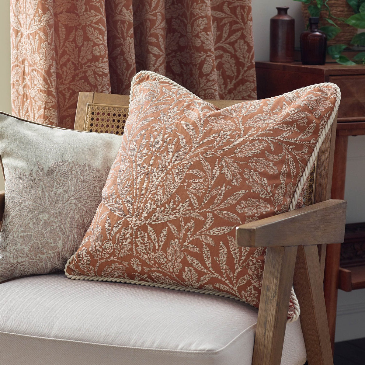 Acorn Tonal Jacquard polyester cushion in brick orange with cream woven acorn-leaf pattern and cord trim, shown on an armchair in a living room setting.