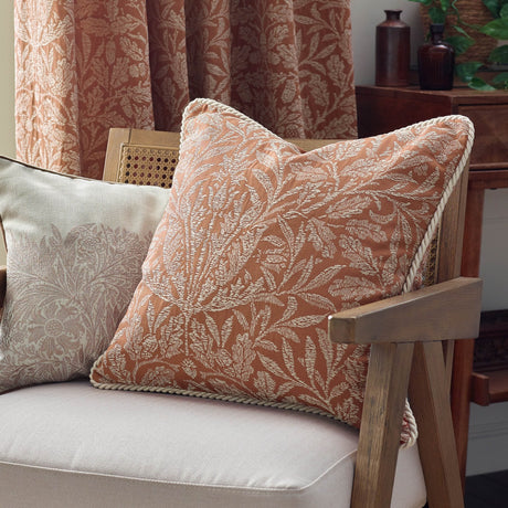 Acorn Tonal Jacquard polyester cushion in brick orange with cream woven acorn-leaf pattern and cord trim, shown on an armchair in a living room setting.