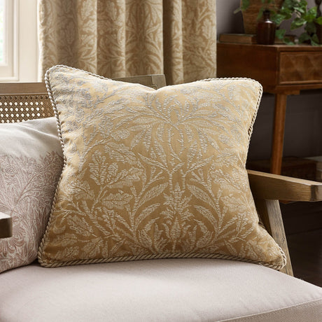 Square gold jacquard cushion with woven cream acorn and foliage pattern and twisted cord trim, shown on a sofa in a living room setting.