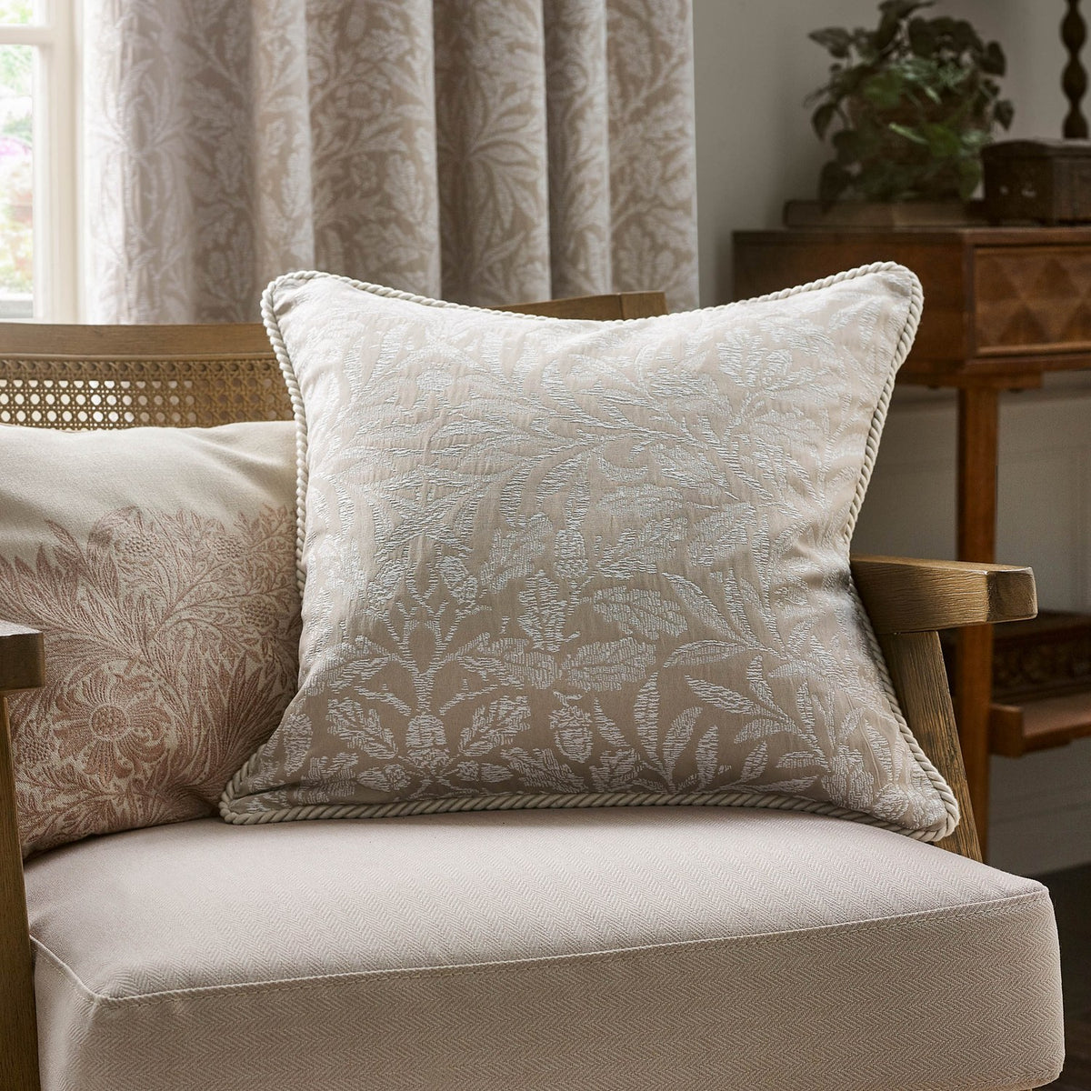 Square Acorn Tonal Jacquard Linen Cushion in cream with woven leaf and acorn pattern and corded edge trim, shown on an armchair in a living room setting.