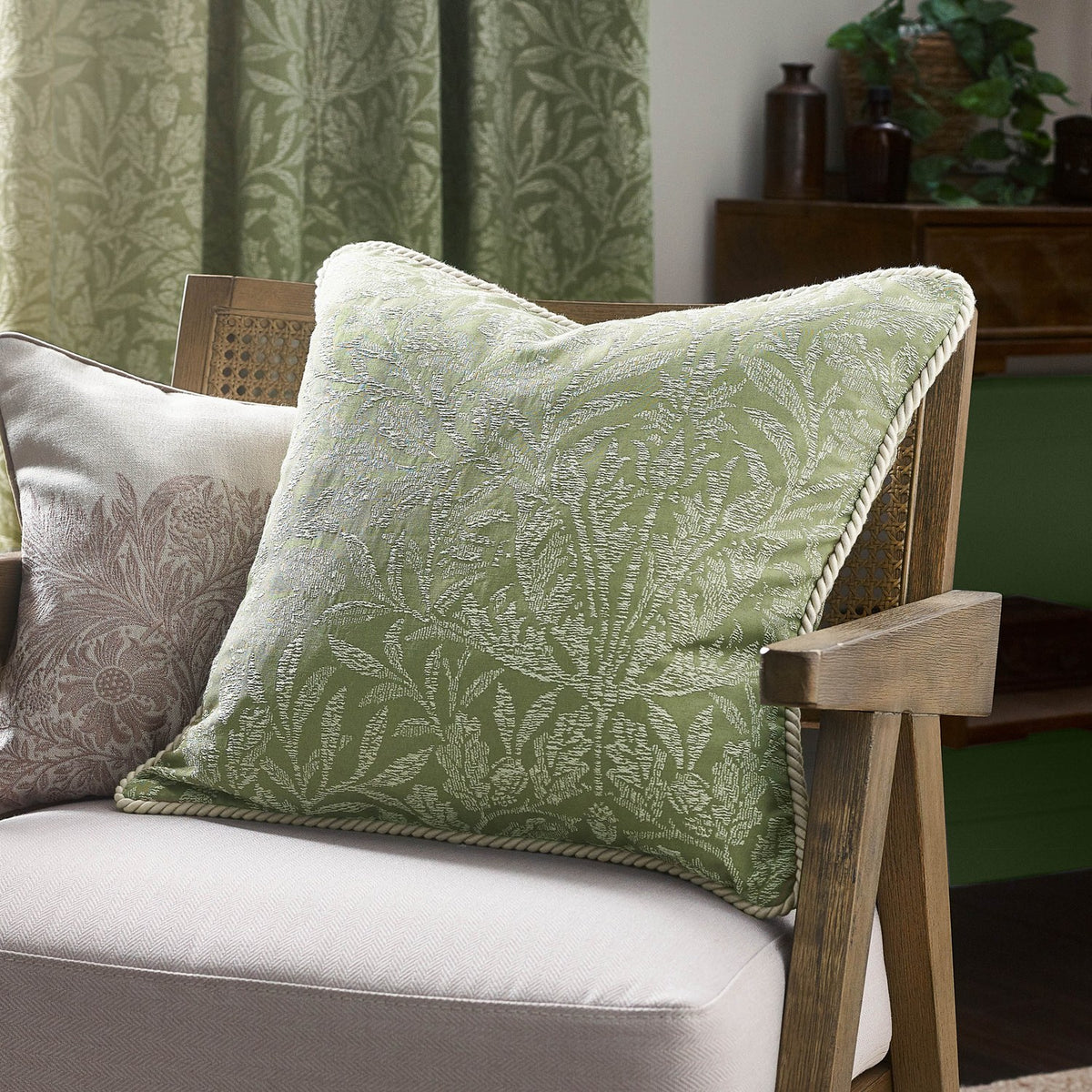 Square sage green Acorn Tonal jacquard cushion with woven acorn branch pattern and cord trim, shown on a cane-armed chair in a living room.