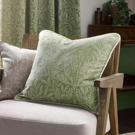 Square sage green Acorn Tonal jacquard cushion with woven acorn branch pattern and cord trim, shown on a cane-armed chair in a living room.