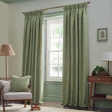 Sage green Acorn Tonal jacquard cushion with leaf branch pattern, styled on a beige armchair in a living room with green curtains.