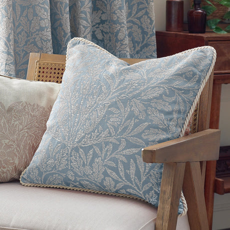 Square Acorn Tonal Jacquard cushion in Wedgewood blue with woven leaf and acorn pattern and cream cord trim, shown on a living room armchair.