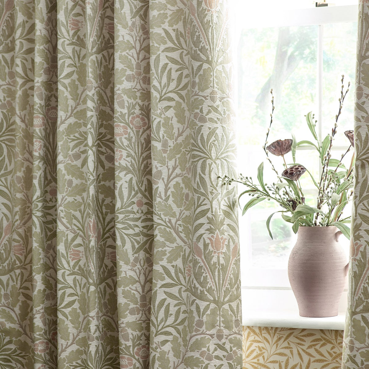 Acorn Multi Jacquard Chenille pencil pleat curtains in stone/sage, woven oak leaf and acorn pattern, shown pencil pleated at a window beside a pink jug vase.