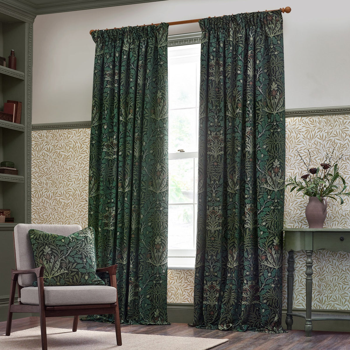 Acorn Multi Jacquard cushion in Thyme/Forest green on a fabric armchair, showing woven oak branch and acorn pattern, in a living room with matching curtains.