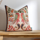 Akamba Animal Linen cushion in multicolour, featuring mirrored parrots in coral and green with jungle florals and paisley border on a natural ground, shown on a wooden surface.