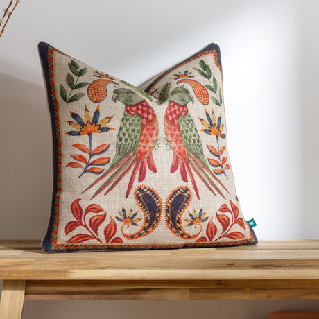 Akamba Animal Linen cushion in multicolour, featuring mirrored parrots in coral and green with jungle florals and paisley border on a natural ground, shown on a wooden surface.