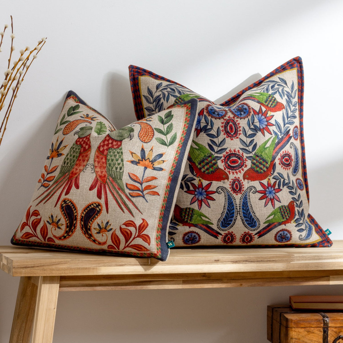 Two Akamba Animal linen cushions with mirrored parrot and paisley prints in red, green and amber, shown on a wooden bench against a light wall.