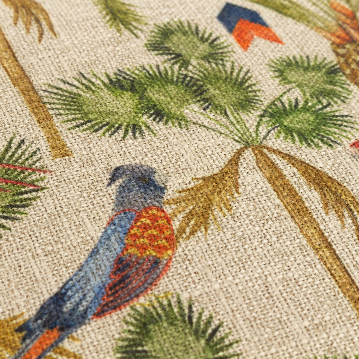 Akamba Animal Linen Cushion in multicolour, close-up of a blue and orange parrot among printed palm trees and foliage on a natural linen-look ground.