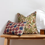 Akamba Animal Linen Cushion in natural linen-look fabric with a printed parrot and palm tree pattern in blue, red, orange and green, shown with a patterned bolster on a wooden bench.