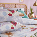 Alchemy Reversible Abstract Duvet Cover Set in cream polycotton, styled on a bed with pillowcases showing sacred hearts, suns, snakes and shells in pink, purple and gold tones.