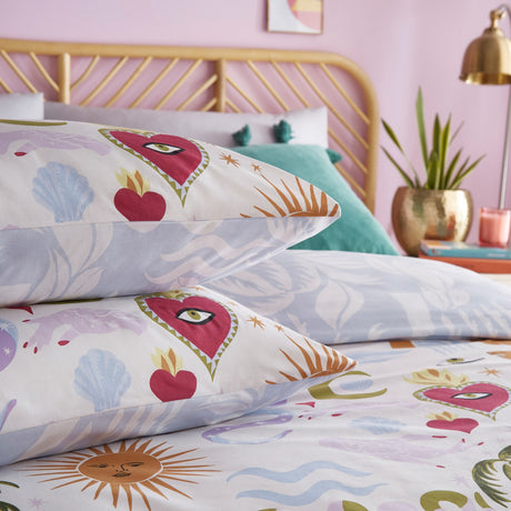 Alchemy Reversible Abstract Duvet Cover Set in cream polycotton, styled on a bed with pillowcases showing sacred hearts, suns, snakes and shells in pink, purple and gold tones.