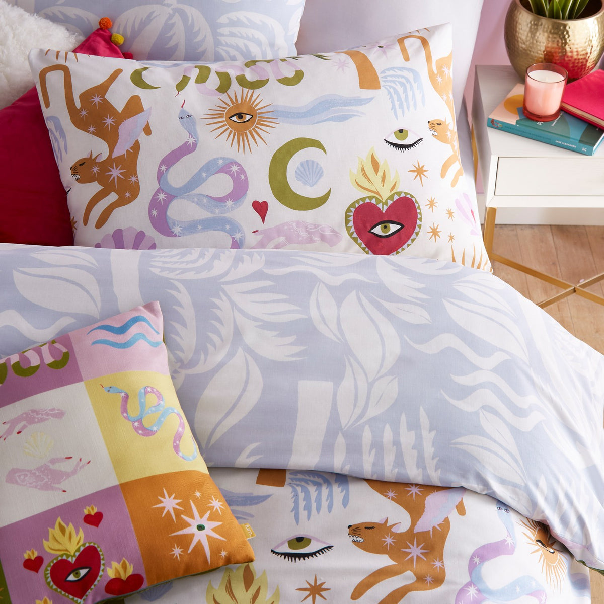 Alchemy reversible duvet cover set on a bed, cream ground printed with suns, moons, snakes, deer and sacred hearts, reverse side in soft blue leaf pattern, styled with matching printed cushions.