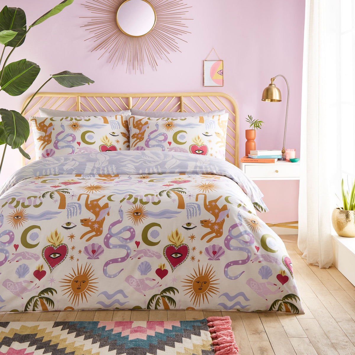 Alchemy Reversible Abstract Duvet Cover Set in Multicolour, styled on a bed in a pink bedroom with cream fabric featuring suns, snakes, hearts, moons and palm motifs, plus matching pillowcases.