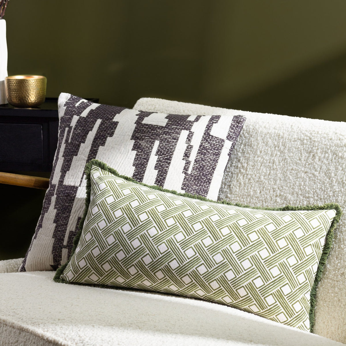 Alexa Reversible Cotton Cushion in olive green with cream basket weave pattern, rectangular bolster shape with fringe trim, styled on a boucle sofa beside a patterned cushion.
