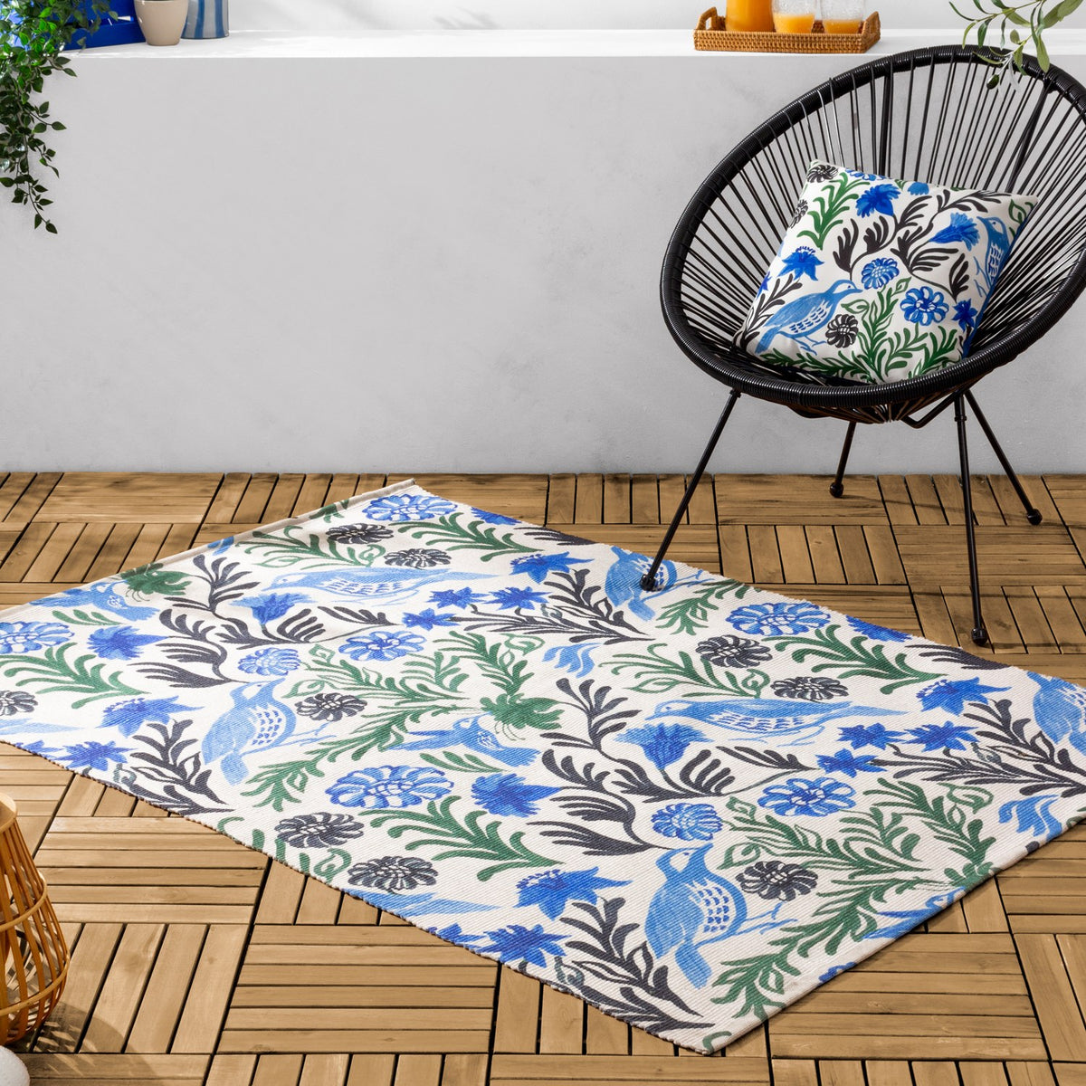 Aljento Abstract Polyester Cushion in blue, green and black floral bird print on a woven black outdoor chair, matching a rug on wooden decking.