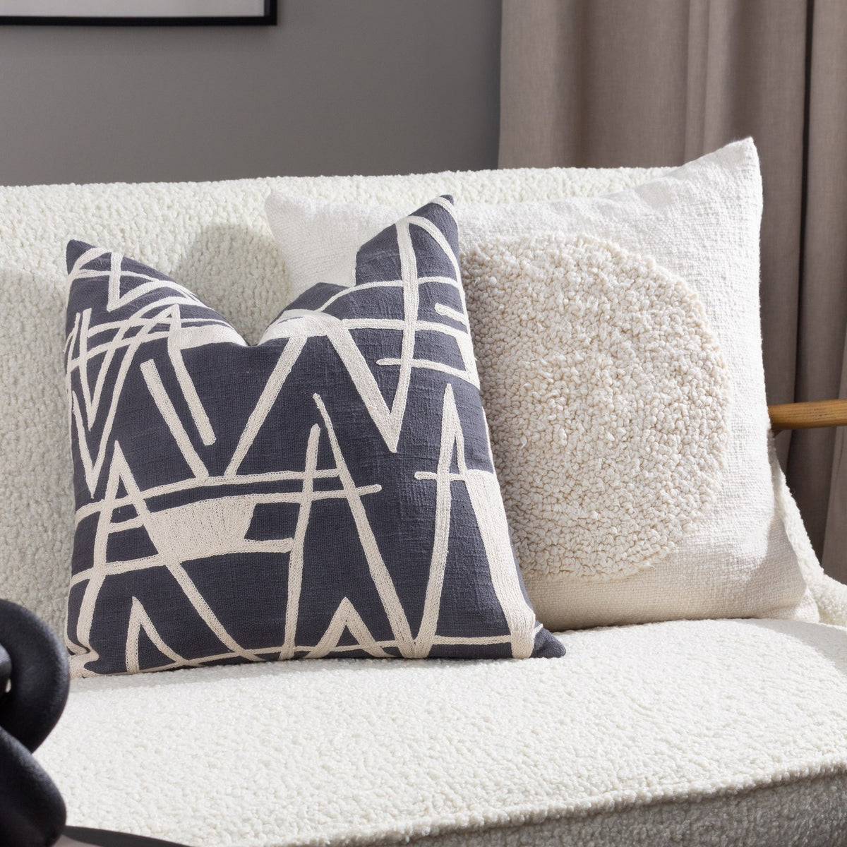 Almo Abstract Cotton Cushion in ecru with a tufted boucle circle on a woven cotton face, shown on a cream boucle sofa beside a patterned grey and cream cushion.