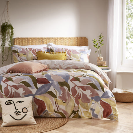 Amarosa Reversible Abstract Duvet Cover Set in Plaster on a bed with rattan headboard, featuring an earthy floral leaf print in clay, olive, blue and gold with coordinating pillows.