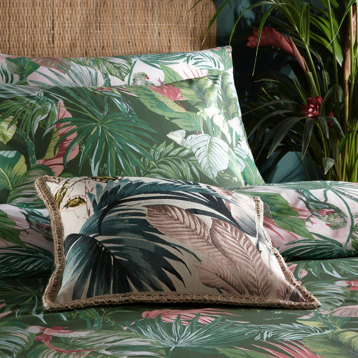 Amazonia Reversible Jungle Duvet Cover Set in Jade, styled on a bed with a matching tropical print cushion, layered pillowcases, and green foliage in the background.