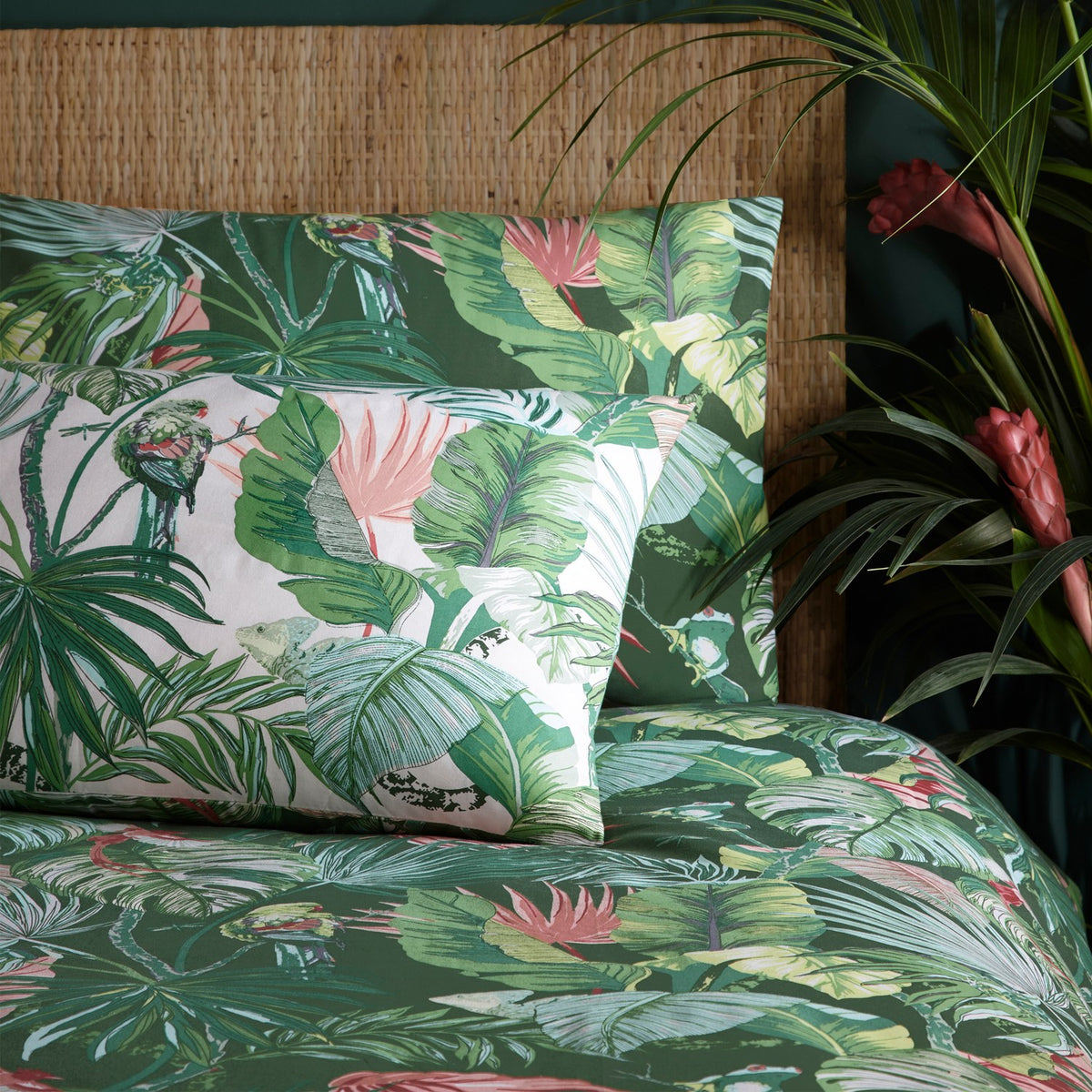 Amazonia Reversible Jungle Duvet Cover Set in Jade, shown on a bed with matching pillowcase featuring green tropical foliage and parrot print, styled against a woven headboard and potted plants.