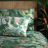Amazonia Reversible Jungle Duvet Cover Set in Jade, shown on a bed with matching pillowcase featuring green tropical foliage and parrot print, styled against a woven headboard and potted plants.