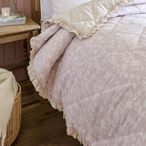 Amelia Ditsy Cotton Bedspread in blush, quilted diamond texture with a ditsy floral print and cream frilled edge, shown on a bed beside a wooden nightstand.