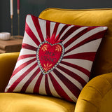 Amor Eterno velvet cushion in red and white, featuring a hand-painted flaming heart with "Amor Eterno" text amid radiating sunburst stripes, piped edge trim, shown on a mustard sofa.