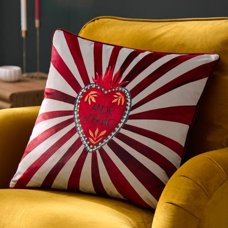 Amor Eterno velvet cushion in red and white, featuring a hand-painted flaming heart with "Amor Eterno" text amid radiating sunburst stripes, piped edge trim, shown on a mustard sofa.