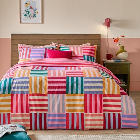 Ana Stripe Reversible Geometric Duvet Cover Set in Multi, patchwork side shown with pink, red, orange, teal, lilac and cream striped blocks, styled on a bed with a wooden headboard.