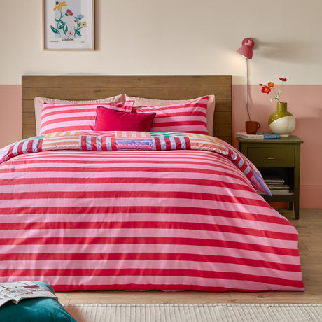 Ana Stripe Reversible Duvet Cover Set on a bed, showing broad pink and red horizontal stripes with a folded corner revealing the multicoloured geometric patchwork reverse.