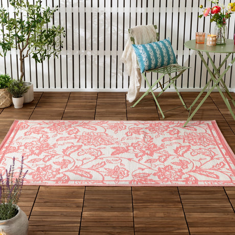 Anemone Jacquard Floral Rug in coral pink and off-white, rectangular with scrolling leaf and floral border, shown on a wooden deck beside a green bistro chair and table.