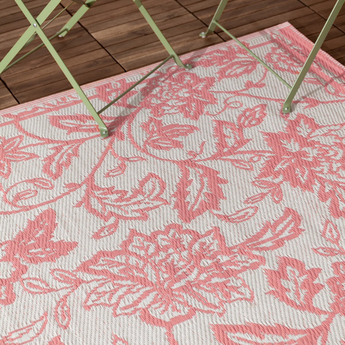Anemone Jacquard Floral Rug in coral pink and off-white, woven Jacobean-style floral pattern, shown outdoors on a wooden deck beneath a folding table's legs.