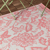 Anemone Jacquard Floral Rug in coral pink and off-white, woven Jacobean-style floral pattern, shown outdoors on a wooden deck beneath a folding table's legs.