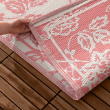 Anemone Jacquard Floral Rug in coral pink and off-white, folded corner revealing woven jacquard texture and bound edge, held by hand over a wooden deck.